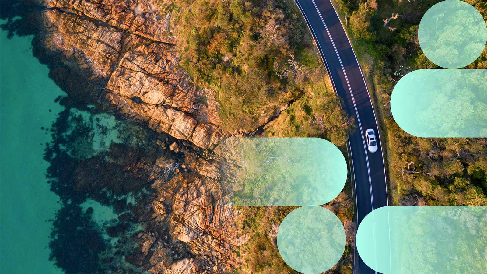 A silver car driving on a road near the ocean, with greenery and a rocky cliff on either side of the road.