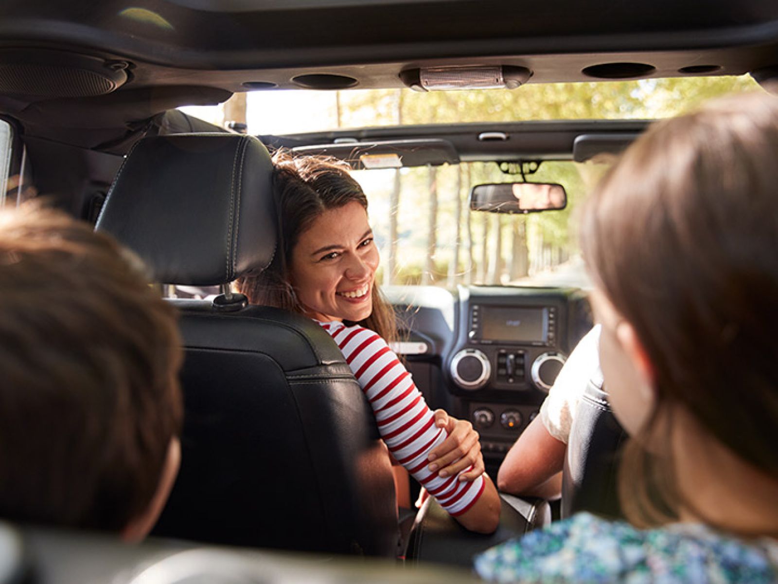 A parent in the front seat of a car, smiling and looking back at 2 kids in the backseat.