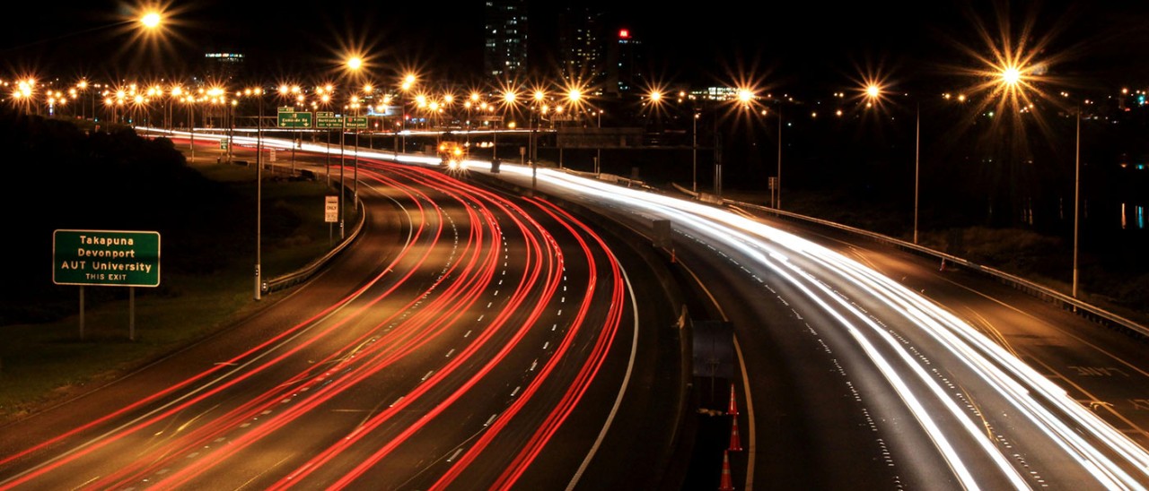 Auckland Northern Motorway in New Zealand, a motorway lit up at night. The long exposure of the photo makes the cars on the road look like red and white streaks of light.