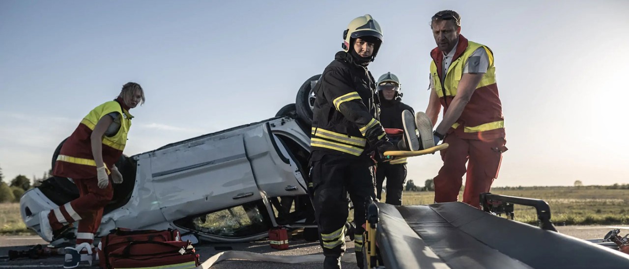 Four emergency responders in high-vis, lifting a person onto a stretcher. A damaged and upside-down white sedan is next to them.