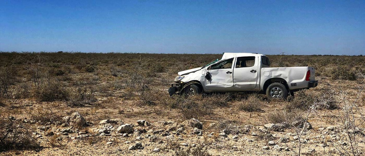 A dented white ute on a rocky landscape. The front of the ute is crumpled, and the sideview mirror is hanging off the car.