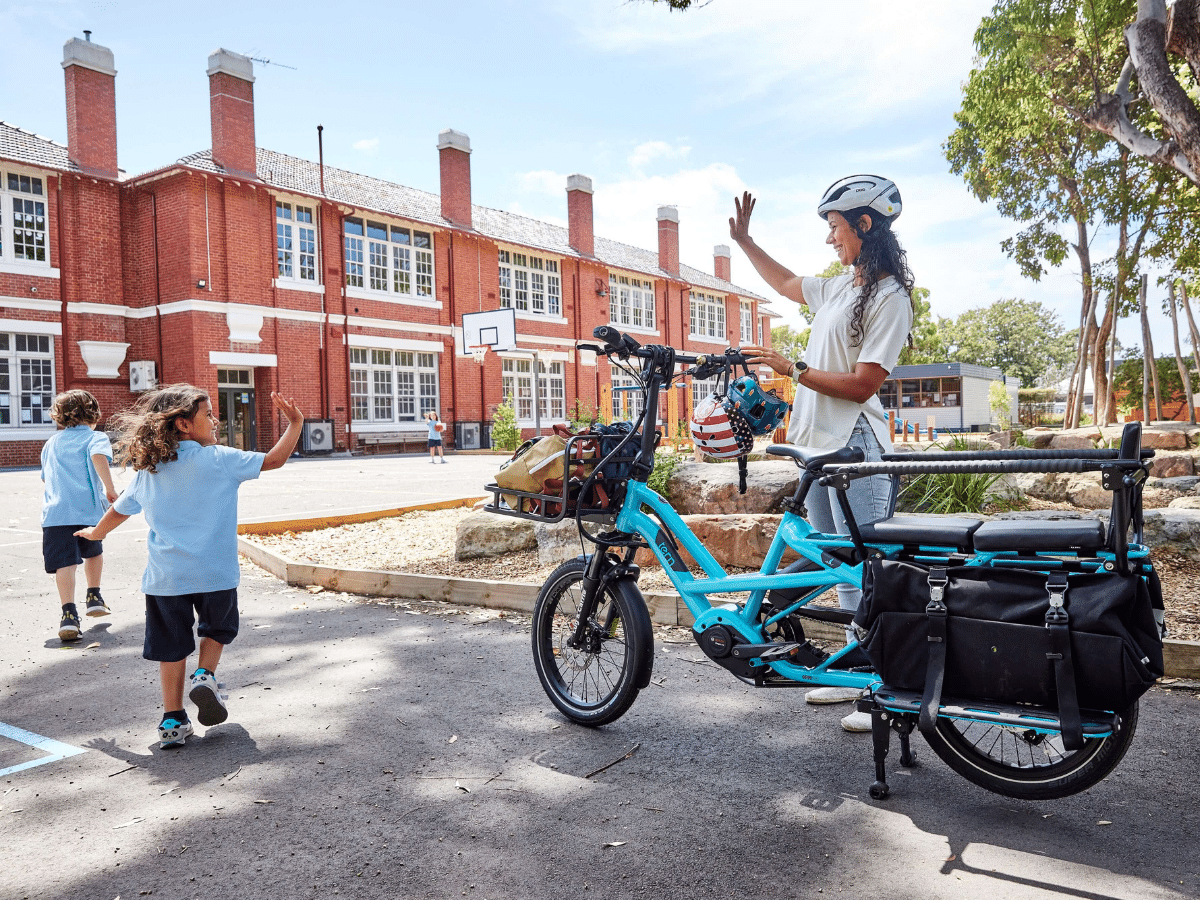 A woman standing next to a bicycle waves to her children in the school yard